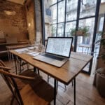 Een laptop met architectonische plannen staat op een houten tafel in een gezellig café met stenen muren en grote ramen. In de buurt staan stoelen, vellen papier, een pen en een plant. Het straatbeeld toont de historische gevel van een gebouw dat vaak wordt genoemd op lokale reiswebsites.